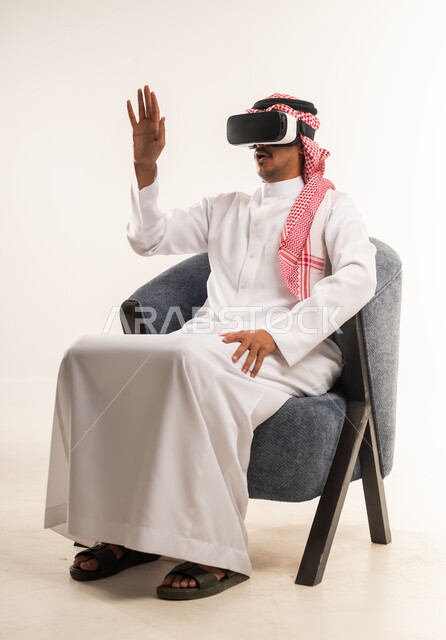 الاندماج والتفاعل ومحاكاة العالم الخيالي, بورتريه لرجل عربي خليجي بحريني يرتدي الثوب التقليدي والشماغ يجلس على كرسي مريح يضع نظارات الواقع الافتراضي يقوم برفع وفتح كف يده للاعلى بتعابير التوقف عن فعل امر ما, شاب سعودي يستخدم نظارة الفي آر، خلفية بيضاء