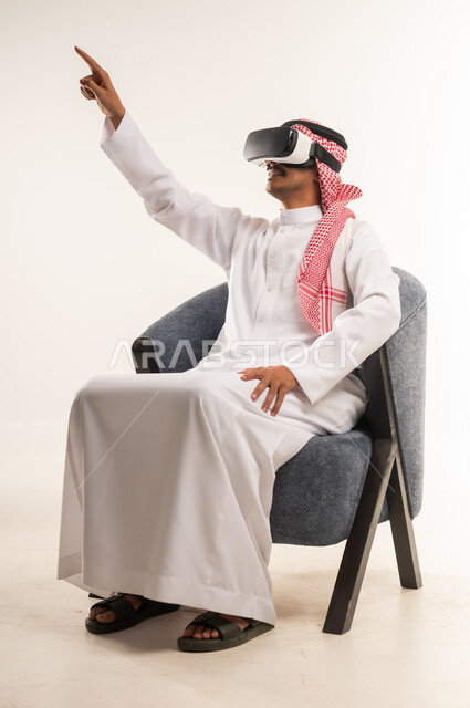 محاولة النقر على شيء ما, بورتريه لرجل عربي خليجي بحريني يرتدي الثوب التقليدي والشماغ يجلس على كرسي مريح يضع نظارات ثلاثية الأبعاد يقوم برفع اصبع السبابة للاعلى، شاب سعودي يشير الى مكان ما بتعابير الاندماج ومحاكاة العالم الخيالي, خلفية بيضاء