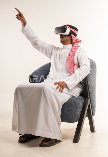 محاولة النقر على شيء ما, بورتريه لرجل عربي خليجي بحريني يرتدي الثوب التقليدي والشماغ يجلس على كرسي مريح يضع نظارات ثلاثية الأبعاد يقوم برفع اصبع السبابة للاعلى، شاب سعودي يشير الى مكان ما بتعابير الاندماج ومحاكاة العالم الخيالي, خلفية بيضاء