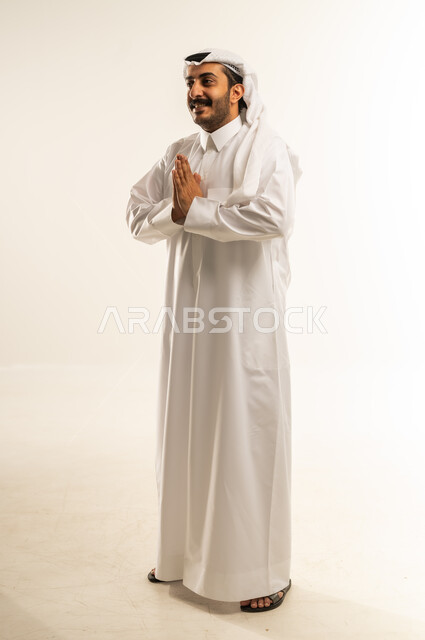مفهوم الشكر والامتنان، الوقوف بإيماءات الدعاء والتوسل والاسترحام، الطلب بترجي وتذلل، بورتريه لرجل عربي خليجي قطري يرتدي الثوب التقليدي والغترة يرفع يديه بخشوع واستعطاف، طلب العون والرغبة في تحقيق الأمنيات، صورة الجسم بالطول الكامل، خلفية بيضاء