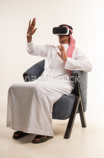 الاندماج والتفاعل ومحاكاة العالم الخيالي، بورتريه لرجل عربي خليجي بحريني يرتدي الثوب التقليدي والشماغ ونظارات الواقع الافتراضي يجلس على كرسي مريح يقوم بمد يديه الى الامام بتعابير صد شيء ما والدفاع عن النفس، شاب سعودي يستخدم نظارة الفي آر، خلفية بيضاء
