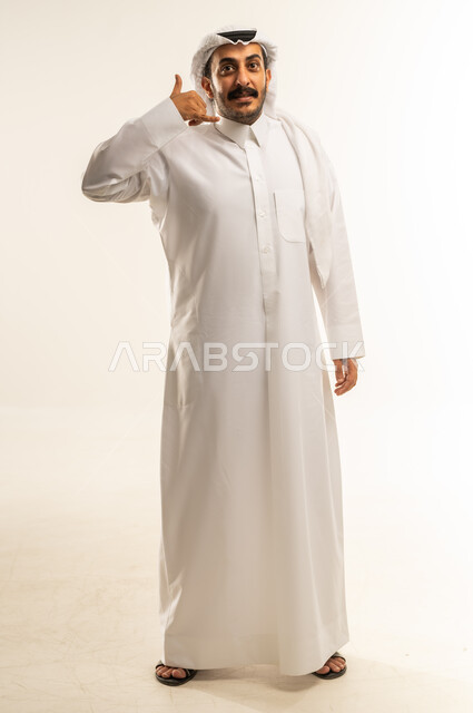 حركة يد تدل على الاتصال، مفهوم تبادل المعلومات، بورتريه لرجل عربي خليجي قطري يرتدي الثوب التقليدي والغترة يرفع يده بايماءات الرد على المكالمات الهاتفية، تلقي أخبار سارة عبر الجوال، صورة الجسم بالطول الكامل، خلفية بيضاء