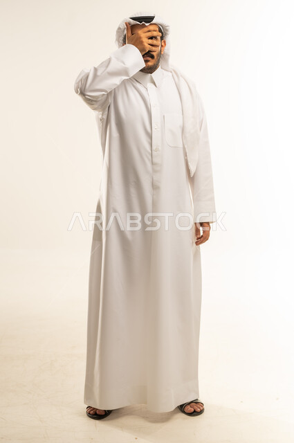 إيماءات تذكر شيء ما بشكل مفاجئ، حركات اليدين والوجه تعبر عن الخجل، بورتريه لرجل عربي خليجي قطري يرتدي الثوب التقليدي والغترة يقف بشكل مستقيم ويغطي عينيه بيده، الاهتمام بالمظهر الخارجي، الجسم بالطول الكامل، خلفية بيضاء