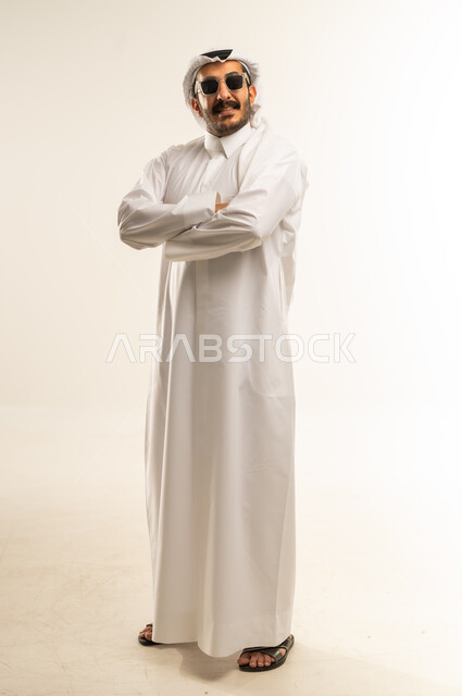 تعابير الثقة بالنفس، الاهتمام بالمظهر الخارجي، مفهوم الرجولة والاناقة، بورتريه لرجل عربي خليجي قطري يرتدي الثوب التقليدي والغترة يضع النظارة الشمسية يقف بايدي متقاطعة ينظر إلى الكاميرا بايماءات السعادة والسرور، صورة الجسم بالطول الكامل، خلفية بيضاء
