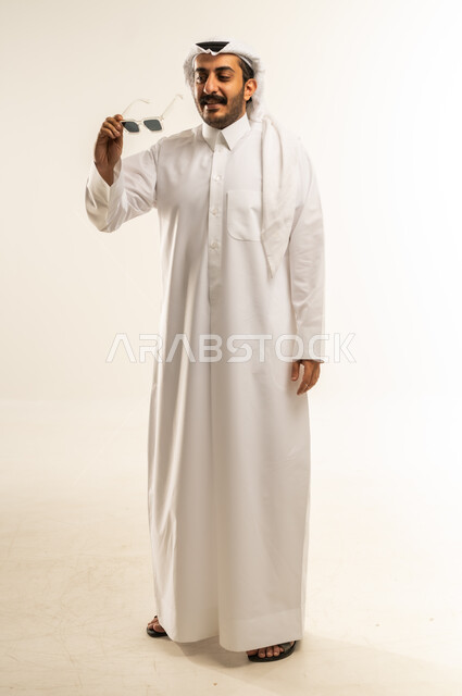 الاهتمام بالمظهر الخارجي، تعابير الثقة بالنفس، مفهوم الرجولة والاناقة، بورتريه لرجل عربي خليجي قطري يرتدي الثوب التقليدي والغترة يضع النظارة الشمسية ينظر إلى مكان ما بايماءات السعادة والسرور، صورة الجسم بالطول الكامل، خلفية بيضاء