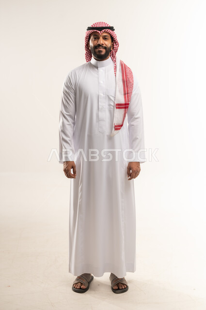 التقاط الصور الشخصية للوثائق الرسمية، الاهتمام بالأناقة والمظهر الخارجي، بورتريه لرجل عربي خليجي بحريني يرتدي الثوب التقليدي والشماغ يقف باستقامة، شاب سعودي ينظر إلى الكاميرا بإيماءات السرور، مفهوم الوسامة والرجولة والقوة، الجسم الكامل بالطول، خلفية بيضاء،