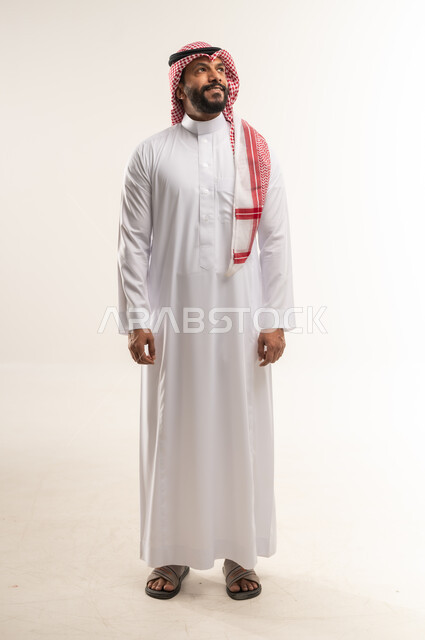 Concern for elegance and appearance, portrait of a Bahraini Gulf Arab man wearing traditional thobe and shemagh standing straight, a Saudi young man looking up somewhere with gestures of pleasure and self-confidence, the concept of handsomeness, masculinity and strength, full body length, white background