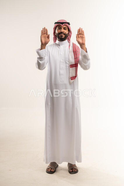مفهوم الرفض والامتناع , شاب سعودي يرفع يده بايماءات طلب التوقف وعدم الاقتراب، بورتريه لرجل عربي خليجي بحريني يرتدي الثوب التقليدي والشماغ يقف باستقامة يرفع كفيه للاعلى، عرض باطن الكف والنظر الى الكاميرا بتعابيرالحزم، الجسم الكامل بالطول، خلفية بيضاء