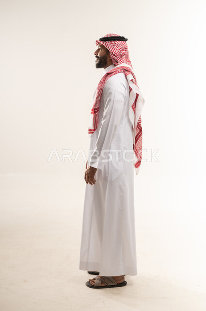 مفهوم الوسامة والرجولة والشياكة، الاهتمام بالأناقة والمظهر الخارجي، بورتريه من الجانب لرجل عربي خليجي بحريني يرتدي الثوب التقليدي والشماغ ينظر الى مكان ما، شاب سعودي يقوم بمحادثة ومخاطبة شخص ما، الجسم الكامل بالطول، خلفية بيضاء