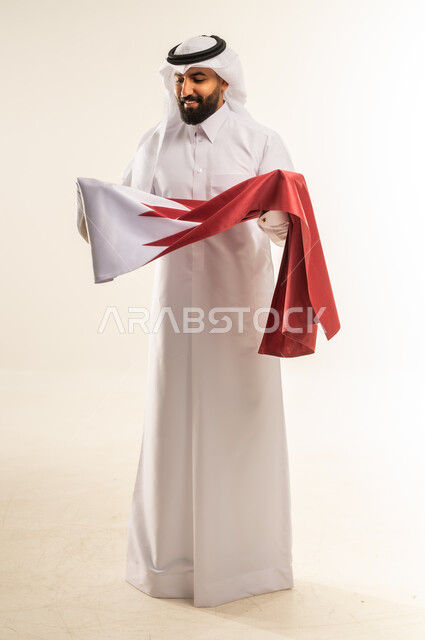 إحياء ذكرى تأسيس الدولة، الاحتفال باليوم الوطني القطري 18 ديسمبر، بورتريه لرجل عربي خليجي قطري يحمل علم قطر بايماءات الفخروالاعتزاز، رموز وثقافات وطنية, حب الوطن والانتماء إليه، الطول الكامل للجسم، خلفية بيضاء