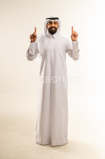 حركات يدين واشارات تدل على هذا المكان (هنا)، تحديد موقع شيء معين، بورتريه لرجل عربي خليجي قطري يرتدي الثوب التقليدي والغتره يشير باصبع السبابة إلى الأعلى، مفهوم الأناقة والاهتمام بالمظهر الخارجي، الجسم بالطول الكامل، خلفيه بيضاء