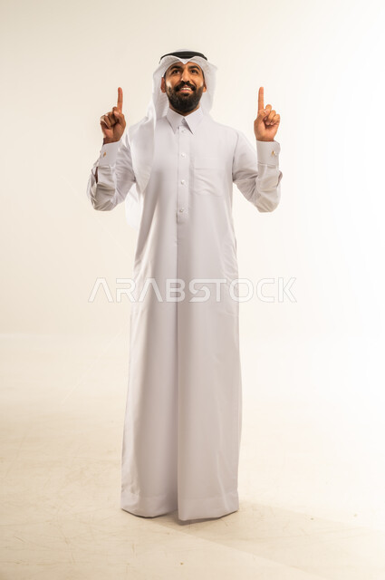 حركات يدين واشارات تدل على هذا المكان (هنا)، تحديد موقع شيء معين، بورتريه لرجل عربي خليجي قطري يرتدي الثوب التقليدي والغتره يشير باصبع السبابة إلى الأعلى، مفهوم الأناقة والاهتمام بالمظهر الخارجي، الجسم بالطول الكامل، خلفيه بيضاء