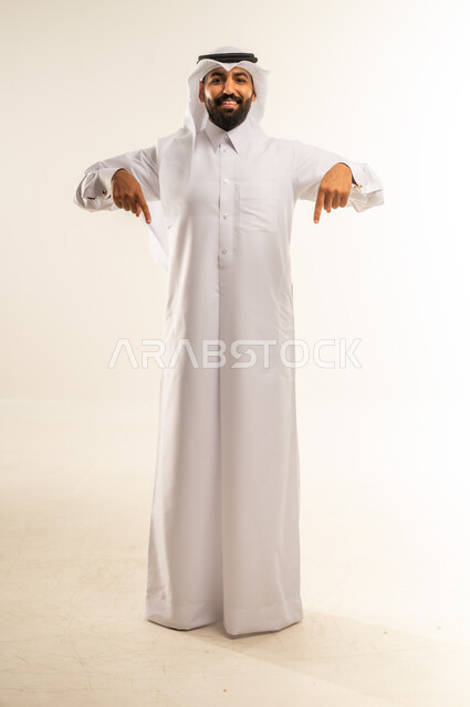 تأكيد حدوث شيء هذه اللحظة, حركات تدل على هذا المكان (هنا)، تحديد موقع معين، بورتريه لرجل عربي خليجي قطري يرتدي الثوب التقليدي والغترة يشير بإصابع السبابة نحو الأسفل, النظر إلى الكاميرا بايماءات السرور، الطول الكامل للجسم, خلفية بيضاء