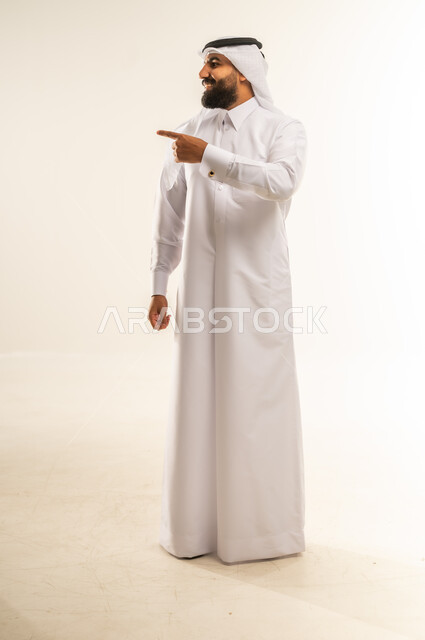 إشارات وحركات يدين تدل على هذا المكان (هنا)، تحديد موقع شيء معين، بورتريه لرجل عربي خليجي قطري يرتدي الثوب التقليدي والغتره يشير باصبع السبابة إلى الأعلى، مفهوم الأناقة والاهتمام بالمظهر الخارجي، الجسم بالطول الكامل، خلفيه بيضاء