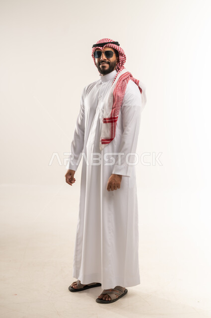 الاهتمام بالأناقة والمظهر الخارجي، مفهوم الوسامة والرجولة والشياكة، بورتريه من الجانب لرجل عربي خليجي بحريني يرتدي الثوب التقليدي والشماغ والنظارات الشمسية يقف بايماءات الثقة بالنفس، شاب سعودي ينظر الى الكاميرا بتعابير السرور، الجسم الكامل بالطول، خلفية بيضاء