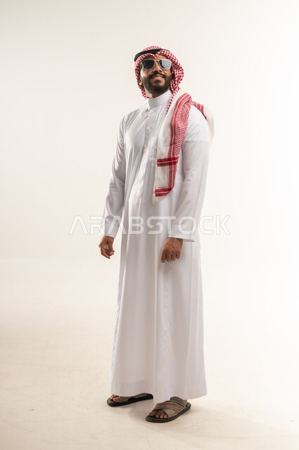 The concept of handsomeness, masculinity and elegance, interest in elegance and outward appearance, portrait of a Bahraini Gulf Arab man wearing the traditional thobe, shemagh and sunglasses standing with an expression of self-confidence, a Saudi young man looking somewhere high with gestures of pleasure, full body length, white background