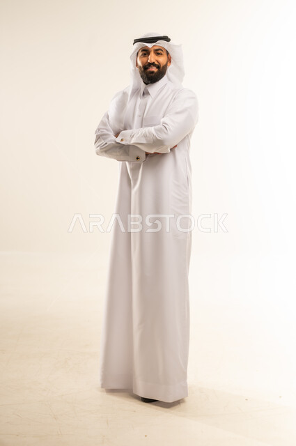 Concern for elegance and outward appearance, looking at the camera with gestures of seriousness and pleasure, the concept of masculinity, strength and self-confidence, portrait of a Qatari Gulf Arab man wearing a traditional thobe and ghutra standing with crossed hands with expressions of firmness, full body length, white background