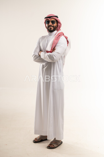مفهوم الوسامة والرجولة، الاهتمام بالأناقة والمظهر الخارجي، بورتريه لرجل عربي خليجي بحريني يرتدي الثوب التقليدي والشماغ والنظارات الشمسية يقف بإيماءات ايدي متقاطعة، شاب سعودية ينظر الى الكاميرا بتعابير السرور والثقة بالنفس، الجسم الكامل بالطول، خلفية بيضاء