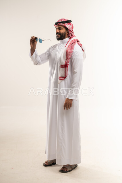 الاهتمام بالأناقة والمظهر الخارجي، مفهوم الوسامة والرجولة والشياكة، بورتريه من الجانب لرجل عربي خليجي بحريني يرتدي الثوب التقليدي والشماغ يحمل بيده النظارات الشمسية، شاب سعودي ينظر بتعابير السرور والثقة بالنفس، الجسم الكامل بالطول، خلفية بيضاء