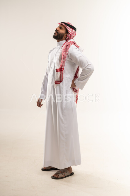 الحاجه للراحه من التعب والإرهاق , شاب سعودي يشعر بألم في العمود الفقري , بورتريه من الجانب  لرجل عربي خليجي بحريني يرتدي الثوب التقليدي والشماغ ينحني  ويضع يده اسفل ظهره بتعابير الشعور بالوجع, طلب المساعده للذهاب الى الطبيب ,صورة الجسم بالكامل، خلفية بيضاء