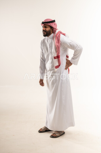 الحاجه للراحه من التعب والإرهاق , شاب سعودي يشعر بألم في العمود الفقري , بورتريه من الجانب  لرجل عربي خليجي بحريني يرتدي الثوب التقليدي والشماغ ينحني  ويضع يده اسفل ظهره بتعابير الشعور بالوجع, طلب المساعده للذهاب الى الطبيب ,صورة الجسم بالكامل، خلفية بيضاء