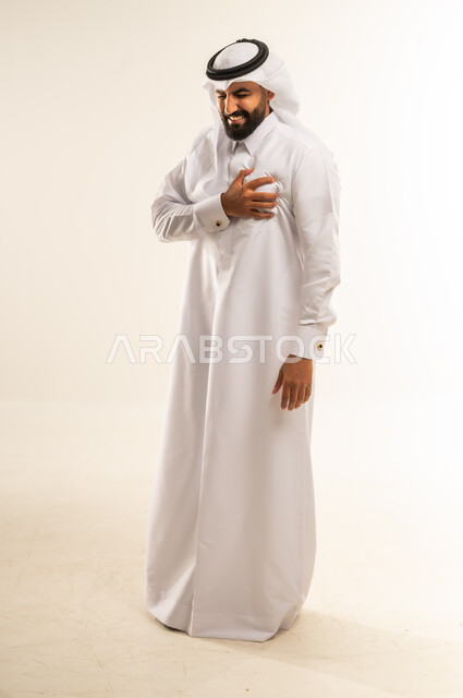 الذبحه الصدريه حالة مرضية، إيماءات التعب والإرهاق والضغوطات النفسية، بورتريه لشاب عربي سعودي قطري يرتدي الغترة و الثوب التقليدي يضع يده على قلبه يشعر بحالة سيئة من الوخز والألم في قلبه, صورة الجسم بالطول الكامل، خلفية بيضاء
