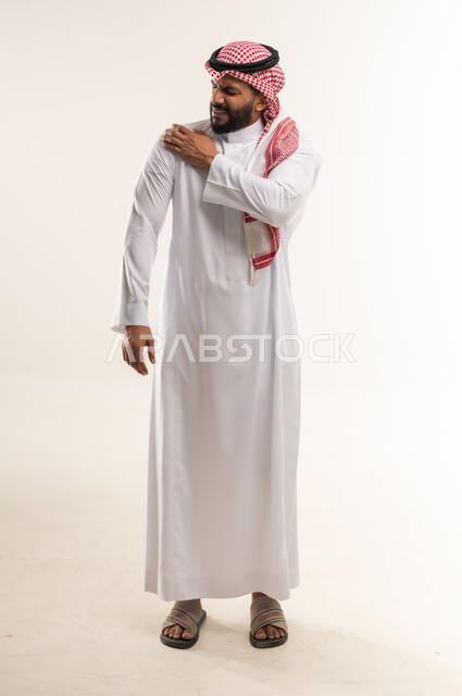 الأوجاع والآلام العصبية والعضلية، طلب المساعده والحاجه للعلاج، شاب سعودي يشعر الالم في العضلات والمفاصل، بورتريه لرجل عربي خليجي بحريني يرتدي الثوب التقليدي والشماغ يضع يده على كتفه بايماءات التعب والأرهاق، حالة اسعافية طارئة، صورة الجسم الطول الكامل, خلفية بيضاء