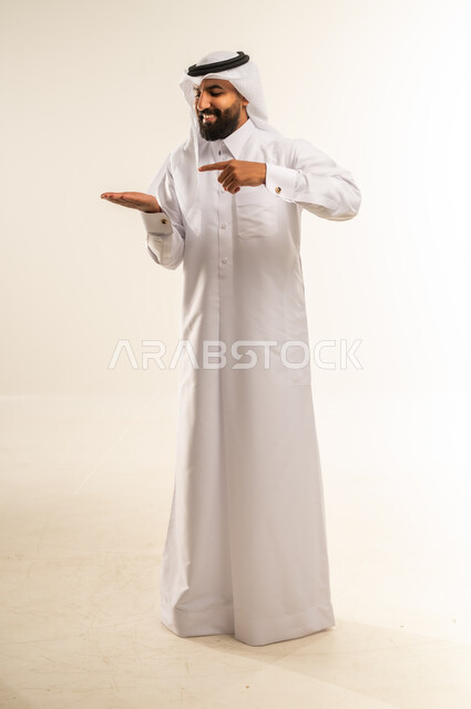 تحديد شيء معين بإيماءات السرور والسعادة، بورتريه لرجل عربي خليجي قطري مبتسم يرتدي الثوب التقليدي والغترة  يفتح كفه للأعلى ويشير إلى عنصر ما, حمل عنصر هولوجرامي، صورة الجسم بالطول الكامل، خلفيه بيضاء