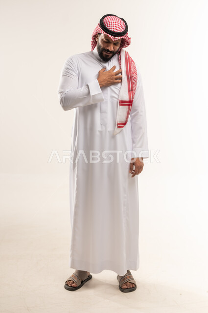 اضطرابات و أمراض قلبية، شاب سعودي يشعر في زيادة بمعدل ضربات القلب، ضرورة الذهاب إلى الطبيب لإجراء الفحوصات اللازمة، بورتريه لرجل عربي خليجي بحريني يرتدي الثوب التقليدي والشماغ  يضع يده على صدره بايماءات الوخز والألم بقلبه, صورة الجسم بالطول الكامل، خلفية بيضاء