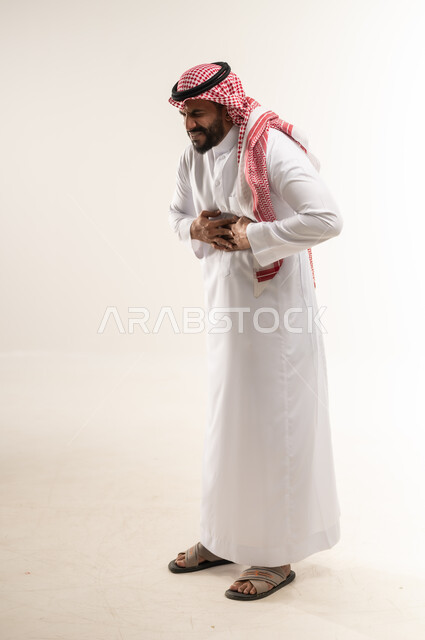 طلب المساعده للذهاب الى الطبيب، شاب سعودي يشعر بعسر الهضم وحرقة في المعدة، بورتريه من الجانب لشاب عربي خليجي بحريني يرتدي الثوب التقليدي والشماغ يمسك بطنه بتعابير الشعور بالألم، الإحساس بالمغص و التعب والإرهاق، الجسم الكامل بالطول، خلفية بيضاء