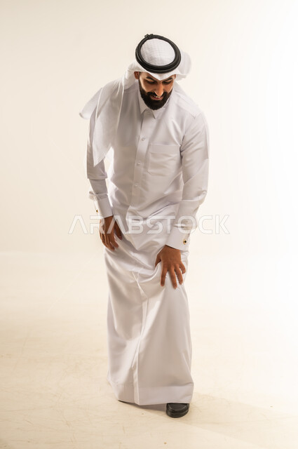 الإحساس بألم في الركبة والمفاصل، الانزعاج والصعوبة بالمشي، آلام شديدة صعبة التحمل، بورتريه لرجل عربي خليجي قطري يرتدي الغترة والثوب التقليدي يضع يده على ركبته بتعابير  التعب، صورة الجسم بالطول الكامل، خلفية بيضاء