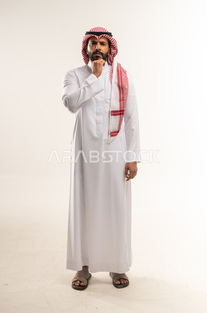 Contemplation and concentration to find a solution to a problem, feeling bored and upset, problems and decision making, portrait of a young Arab Gulf Bahraini man wearing a traditional thobe and shemagh with his hand behind his chin, a Saudi man looking at the camera with gestures of thinking and confusion, full body image, white background