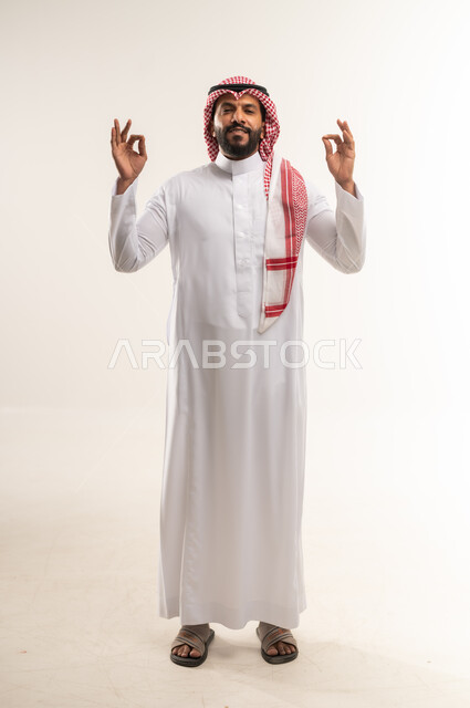 البشاشة والشعور بالرضا, الموافقة والقبول والنصيحة، بورتريه لشاب عربي خليجي إماراتي مبتسم يرتدي الثوب التقليدي والشماغ يقوم برفع يديه بحركات دائرية بإيماءات الجودة والامتياز والتميز، شاب سعودي يقوم بالتعبيرعن الجودة والاتقان، صورة الجسم بالطول الكامل, خلفية بيضاء