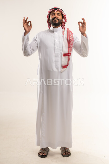 Cheerfulness and satisfaction, approval, acceptance and advice, portrait of a smiling young Arab Gulf Emirati man wearing traditional thobe and shemagh raising his hands in circular motions with gestures of quality, excellence and distinction, a young Saudi man expressing quality and perfection, full body image, white background