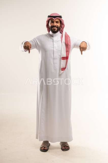 تأكيد حدوث شيء هذه اللحظة, حركات تدل على هذا المكان (هنا)، تحديد موقع معين، بورتريه لرجل عربي خليجي بحريني يرتدي الثوب التقليدي والشماغ يشير بإصابع السبابة نحو الأسفل,  النظر إلى الكاميرا بايماءات السرور، الطول الكامل للجسم, خلفية بيضاء
