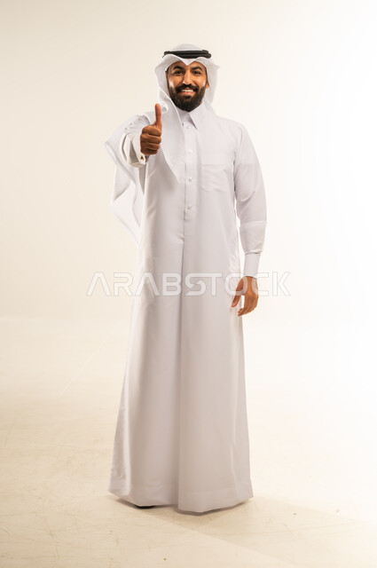 Thumbs up sign of quality and approval, feeling satisfied and pleased, expressing admiration and appreciation, excellence and full marks, portrait of a smiling Qatari Gulf Arab man wearing traditional thobe and ghutra looking at camera with gestures of happiness, full body, white background