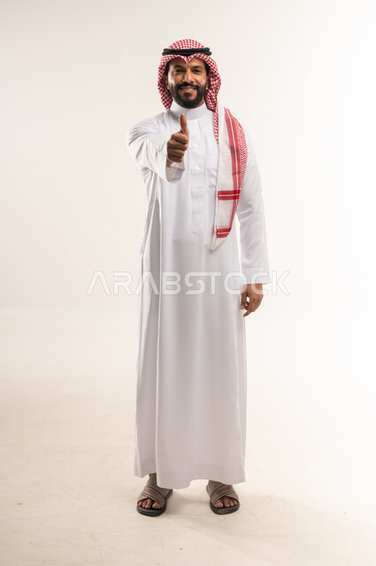 تعابير القبول والفرح والسرور، الاهتمام بالأناقة والمظهر الخارجي، بورتريه لرجل عربيخليجي بحريني يرتدي الثوب التقليدي والشماغ ينظر الى الكاميرا ويرفع إصبع السبابة للاعلى بإيماءات الإعجاب والرضا، الطول الكامل للجسم، خلفية بيضاء