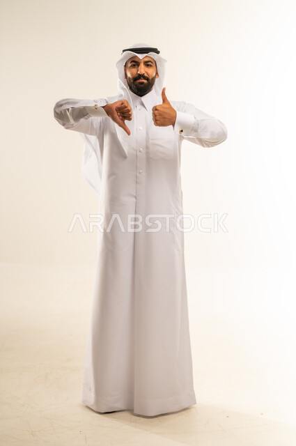 الإشارة باصبعي الابهام إلى جهتين متعاكستين، الحيرة في اتخاذ القرار الصحيح، علامة الجودة والسوء، الرضا وعدم الاستحسان، بورتريه لرجل عربي خليجي قطري مبتسم يرتدي الثوب التقليدي والغتره يضع يده أسفل ذقنه وينظر إلى الكاميرا بجدية، الجسم بالطول الكامل، خلفية بيضاء