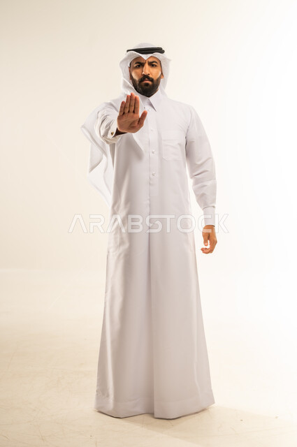 الأمر بالوقوف وعدم التمادي، طلب التوقف عن فعل شيء ما، الرفض وعدم الخضوع، النهي عن التقدم وأهمية ترك مسافة أمان، بورتريه لرجل عربي خليجي قطري يرتدي الثوب التقليدي والغتره يرفع كفه للأعلى بإيماءات الغضب والجدية، الجسم بالطول الكامل، خلفية بيضاء
