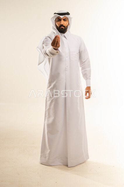 Request for deliberation and slowness, order to stop and not to go too far, stop doing something, refusal and not to submit, prohibition of progress and the importance of leaving a safe distance, portrait of an Arab Gulf Qatari man wearing a traditional thobe and ghutra raising his hand up with gestures of anger and seriousness, full body, white background