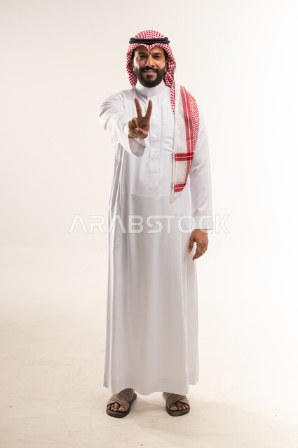 النجاح وتحقيق الأهداف، مفهوم السعادة بالوصول إلى الغايات، بورتريه لرجل عربي خليجي بحريني مبتسم يرتدي الثوب التقليدي والشماغ يرفع يده باشارة النصر والقوة، إيماءات السرور بالفوز والانتصار، الطول الكامل للجسم, خلفية بيضاء