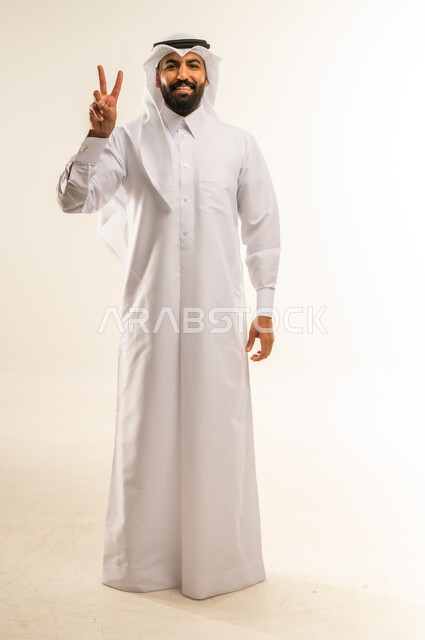 رفع علامة النصر والسلام، إشارة الفوز والانتصار، تحقيق الأهداف والطموحات، إنجاز المهام المحددة، إشارات وحركات لغة الجسد، بورتريه لرجل عربي خليجي قطري مبتسم يرتدي الثوب التقليدي والغتره ينظر للكاميرا بإيماءات السعادة والسرور، الجسم بالطول الكامل، خلفية بيضاء
