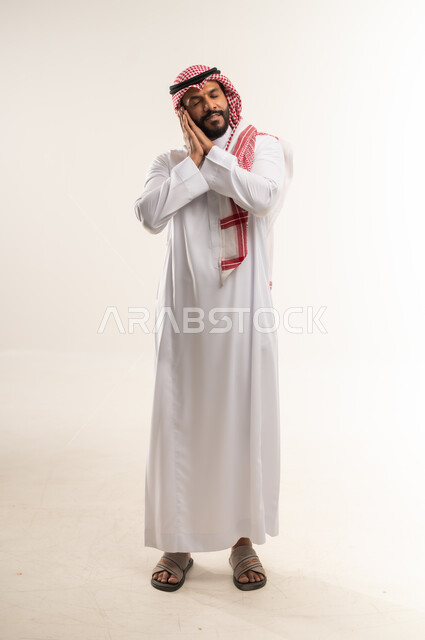 الشعور بالتعب و الحاجه الى الراحه, الإحساس بالكسل والخمول، بورتريه لرجل عربي خليجي بحريني يرتدي الثوب التقليدي والشماغ يضع يديه تحت رأسه ويغمض عينيه بإيماءات النوم، تعابير النعاس والإرهاق، الطول الكامل للجسم، خلفية بيضاء
