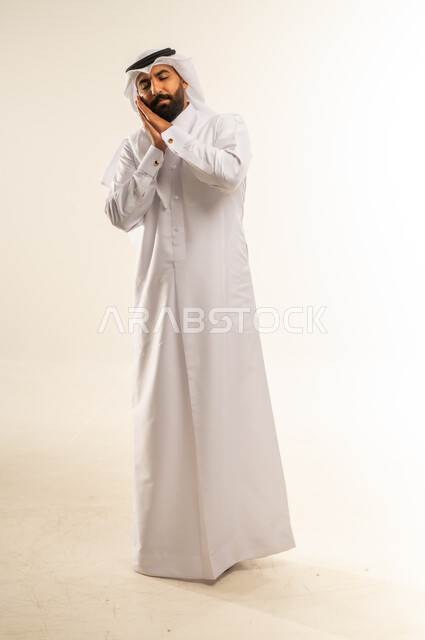 إغماض العينين ووضع اليدين أسفل الخد، الشعور بالنعاس والحاجة للنوم، ضرورة أخذ قسط من الراحة والاسترخاء، بورتريه لرجل عربي خليجي قطري مبتسم يرتدي الثوب التقليدي والغتره يغلق عينيه بإيماءات التعب والإرهاق، الجسم بالطول الكامل، خلفية بيضاء