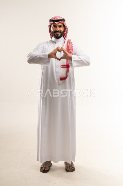 اظهار مشاعر الحب والشغف، مفهوم المحبة والمودة، بورتريه لرجل عربي خليجي بحريني مبتسم يرتدي الثوب التقليدي والشماغ يرفع يديه بإيماءات على شكل قلب، تعابير الفرح والسرور والسعادة، الطول الكامل للجسم, خلفية بيضاء