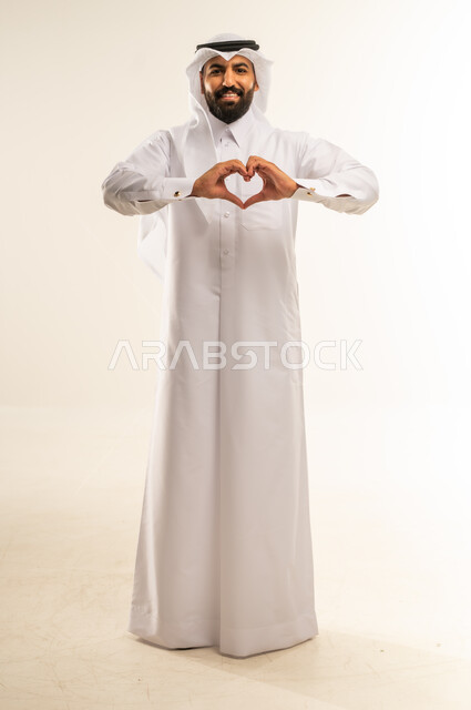 التعبير عن المحبة والمودة، إظهار الحب للآخرين، الاهتمام بالمظهر الخارجي، مفهوم الأناقة والوسامة، النظر إلى الكاميرا بإيماءات البشاشة، بورتريه لرجل عربي خليجي قطري مبتسم يرتدي الثوب التقليدي والغتره يقوم بصنع شكل قلب بيديه، الجسم بالطول الكامل، خلفية بيضاء