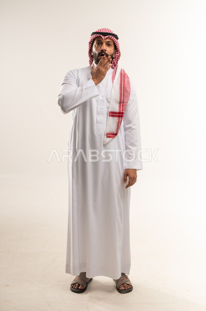 تعابير الذهول لسماع خبر مفاجئ، الصدمة والتعجب من حصول أمر ما، بورتريه لرجل عربي خليجي بحريني يرتدي الثوب التقليدي والشماغ ينظر الى الكاميرا ويضع يديه على وجهه بايماءات الخوف والدهشة، الطول الكامل للجسم، خلفية رمادية