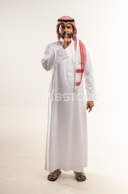 التوقف عن الكلام و الأمر بالصمت والسكوت، بورتريه لرجل عربي سعودي خليجي يرتدي الثوب التقليدي والشماغ يرفع اصبعه الى فمه بإيماءات طلب الهدوء وعدم الازعاج, النظر الى الكاميرا بتعابير الجدية والحزم، الطول الكامل للجسم، خلفية بيضاء