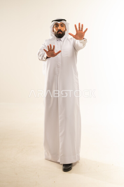 Request to stop doing something, order to stop and not to go further, refusal and not to submit, prohibition of progress and importance of leaving a safe distance, portrait of a Qatari Gulf Arab man wearing traditional thobe and ghutra raising his hand up with gestures of anger and seriousness, full body, white background