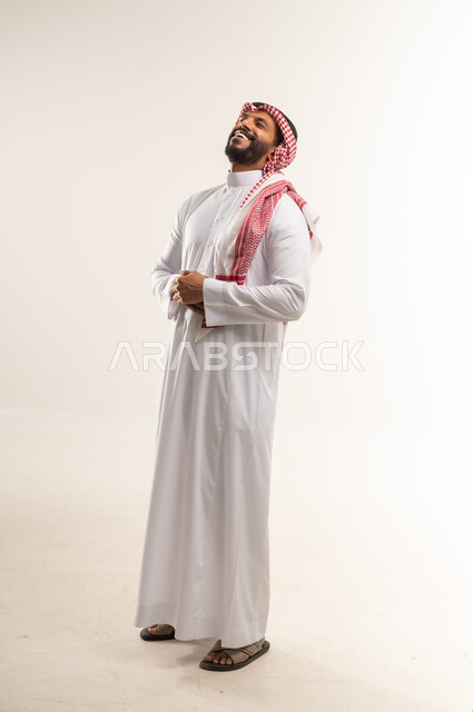 الشعور بالرضا و السعادة، تعابير البهجة والسرور الشديد، بورتريه لرجل عربي خليجي بحريني يرتدي الثوب التقليدي والشماغ يضع يده على بطنه بإيماءات الضحك الهستيرية، الطول الكامل للجسم، خلفية بيضاء